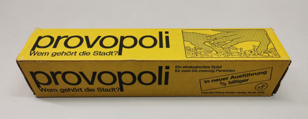 Ein länglicher rechteckiger Pappkarton in leuchtendem Gelb als Verpackung eines Gesellschaftsspiels, bedruckt in schwarzer Schrift u.a. mit Titel des Spiels "provopoli"