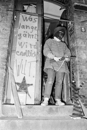 vermummte Person mit Gewehartrappe aus Pappe steht im Hausingang neben Transparent "Was lange gährt wird endlich Wut"