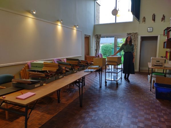 Blick in einen großen Raum mit einem langen Tisch, auf dem Aktenstapel liegen. Am Ende des Tisches steht eine Person neben einem Aktentransportwagen